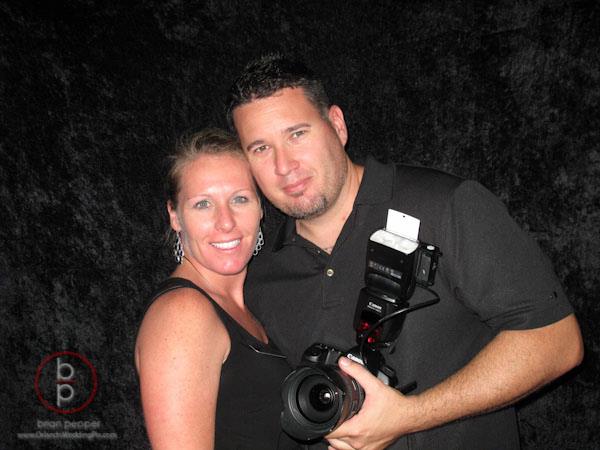 A man and woman stand together against a dark backdrop. The man is holding a camera with a flash attachment. Both are wearing black outfits.