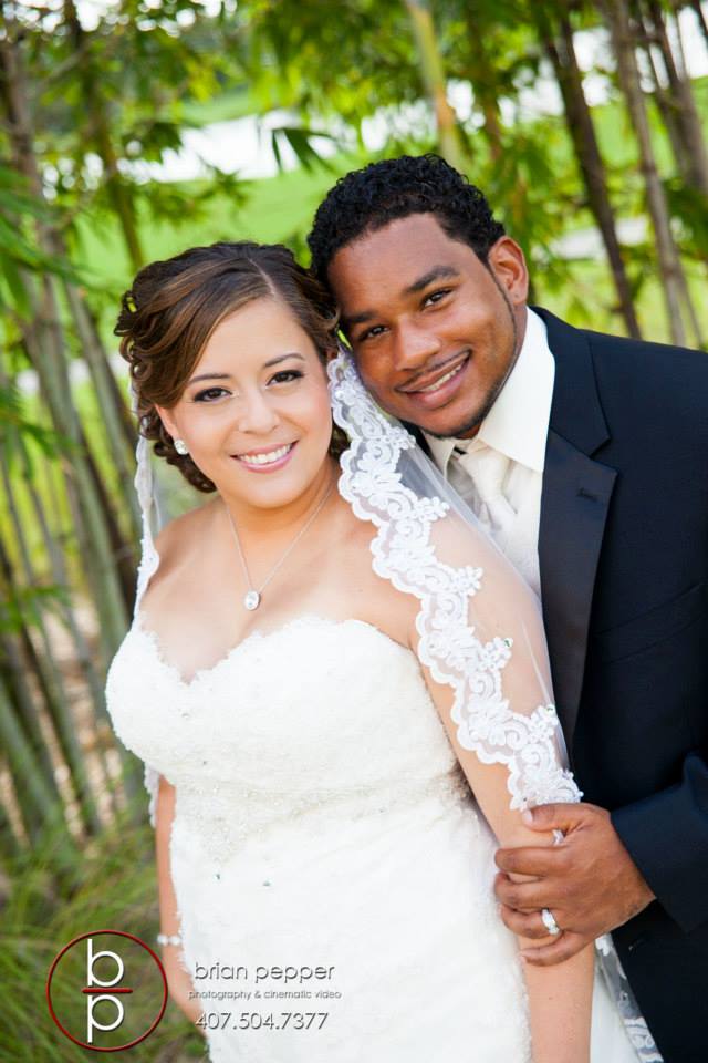 Bride and groom smiling in a garden setting, with the groom in a suit and the bride wearing a white wedding dress with a lace veil.