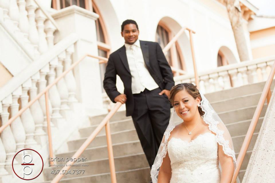 Bride and groom pose on outdoor stairs in formal attire, with the bride in a white dress and veil and the groom in a black suit with a white vest.